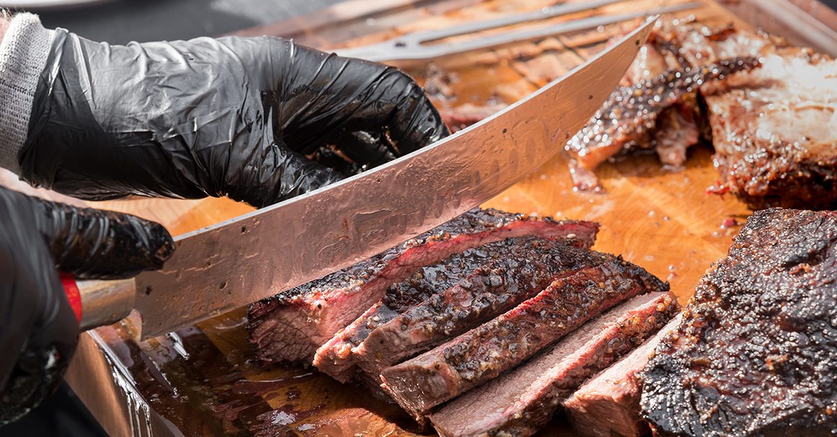 Person in gloves with a large knife cutting beef brisket